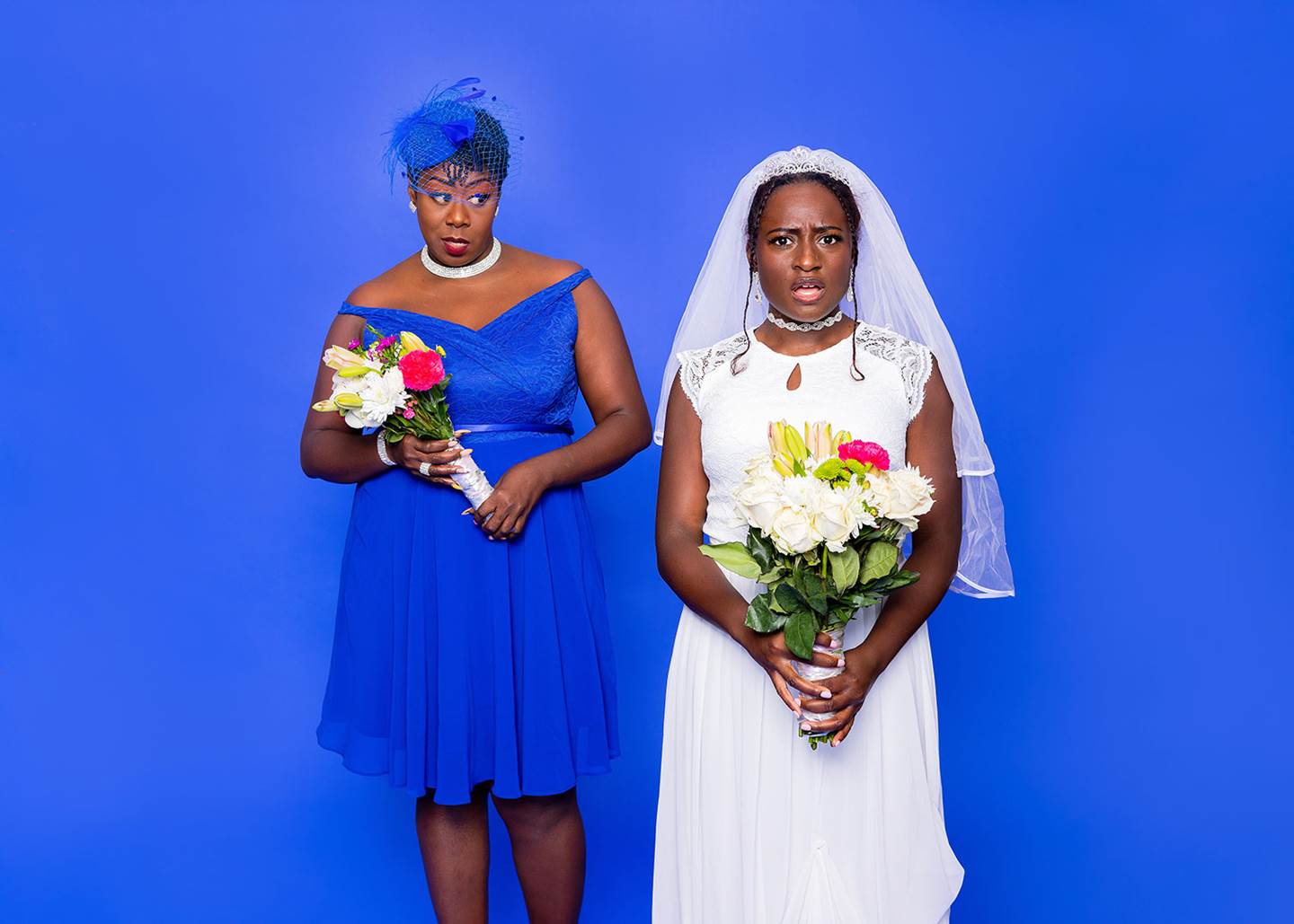 A bride and a bridesmaid wearing their dresses in front of a blue background.